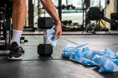 Sportler greift mit einem Disinfektionstuch nach der Hantel danaben befindet sich ein Haufen an verbrauchten Desinfektionstücher am Boden